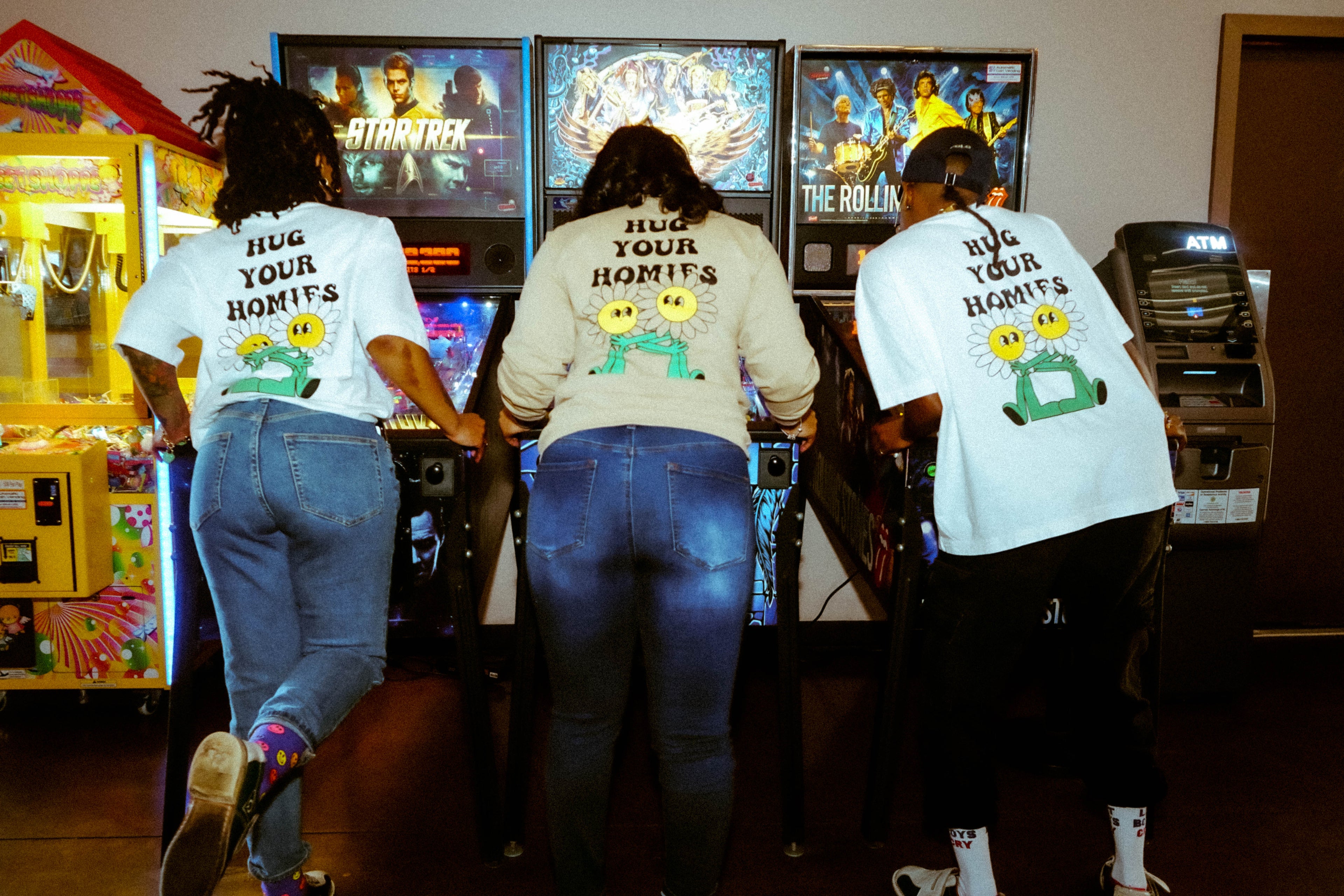 Three friends at an arcade playing games with their backs turned The hug your homies t-shirt by Not a Bad Life.