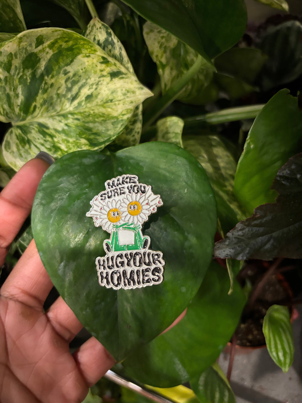 Hug your homies enamel pin laying on a green plant