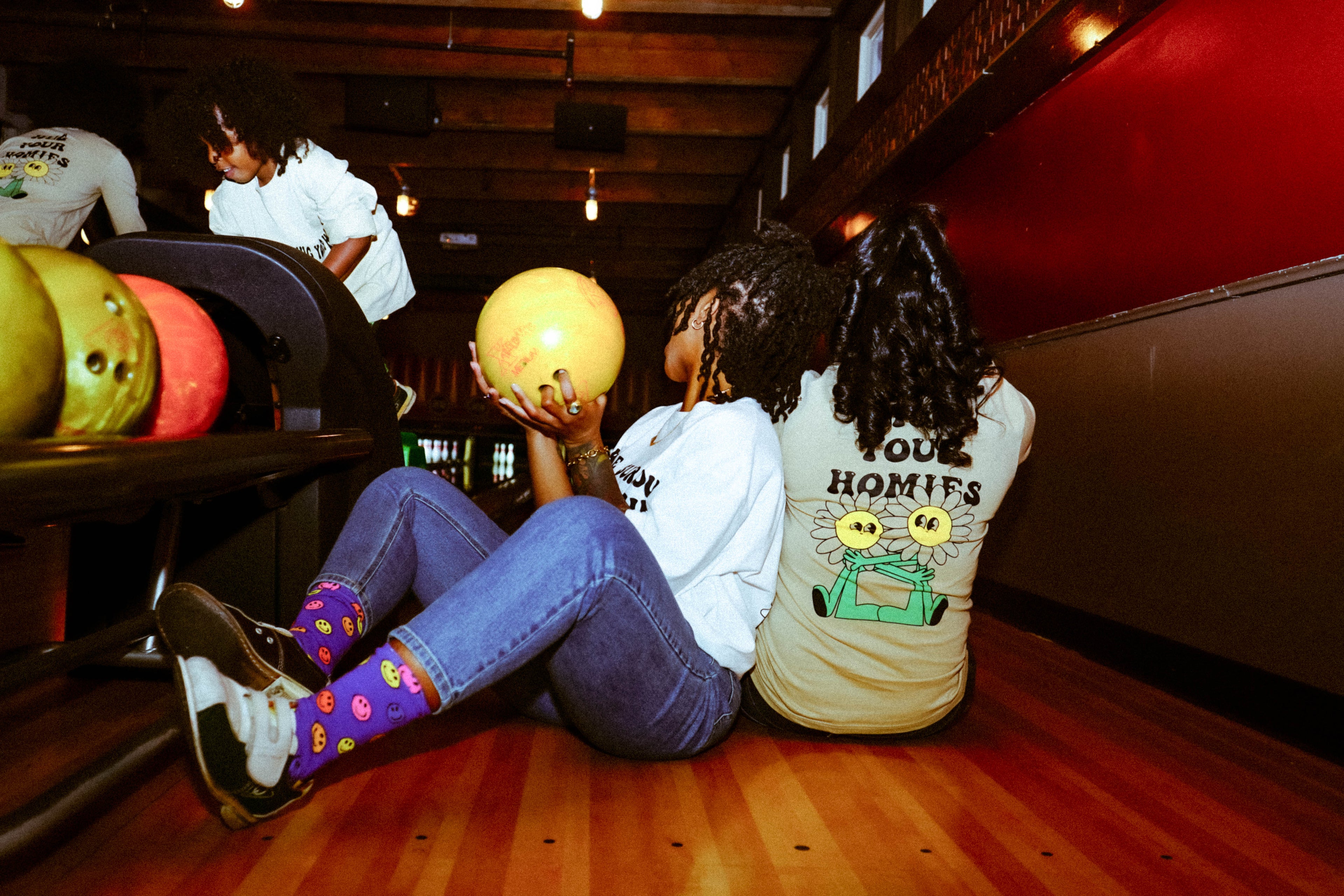 Two girls at the bowling alley leaning on each other wearing hug Your Homies shirts 